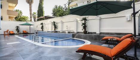 Una piscina con sillas de descanso y sombrillas, rodeada por un muro blanco y palmeras.