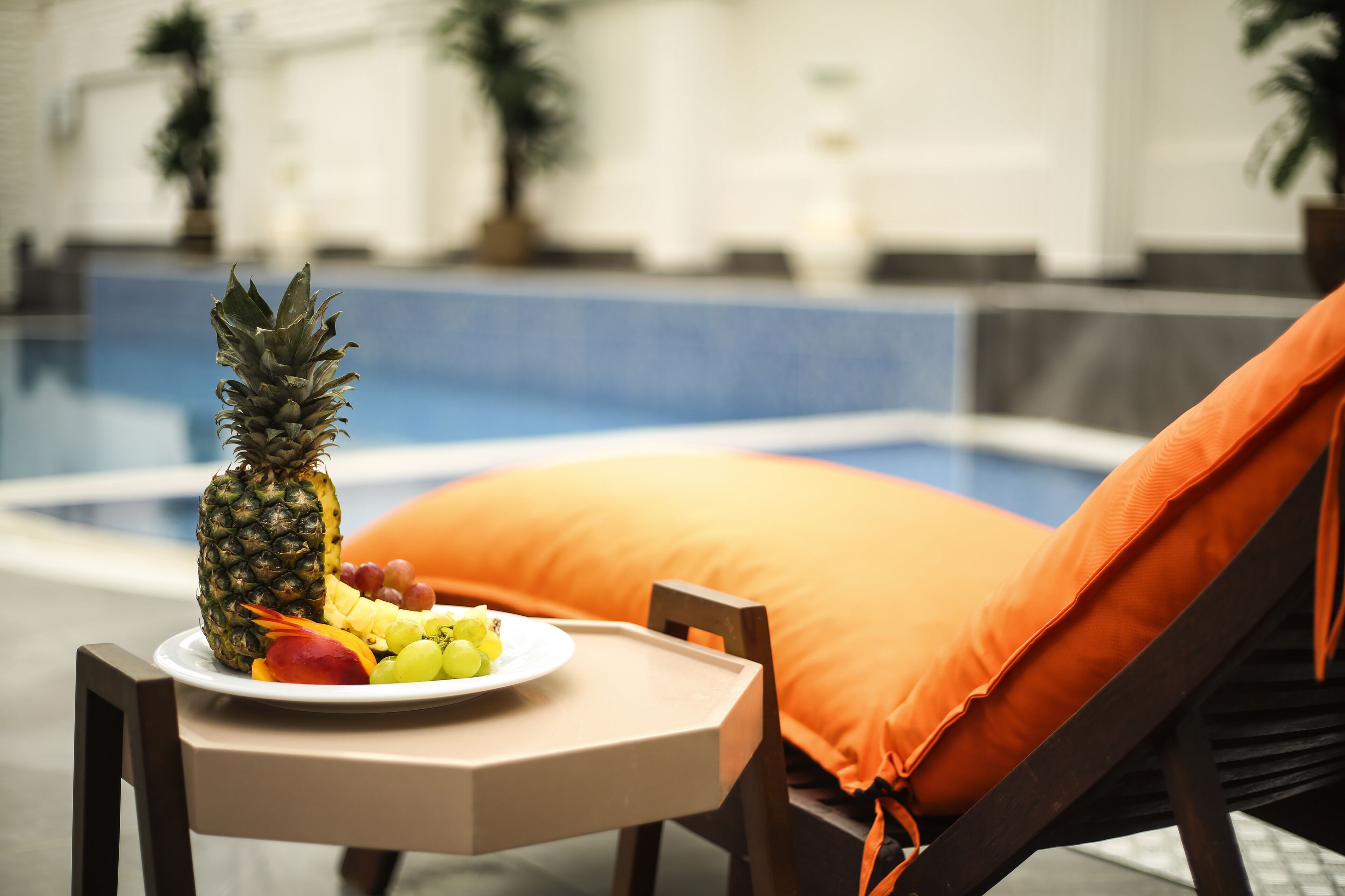 A poolside setting with a table holding a plate of fruit and a pineapple, a lounge chair with an orange cushion, and a blurred background with a pool and potted plants.