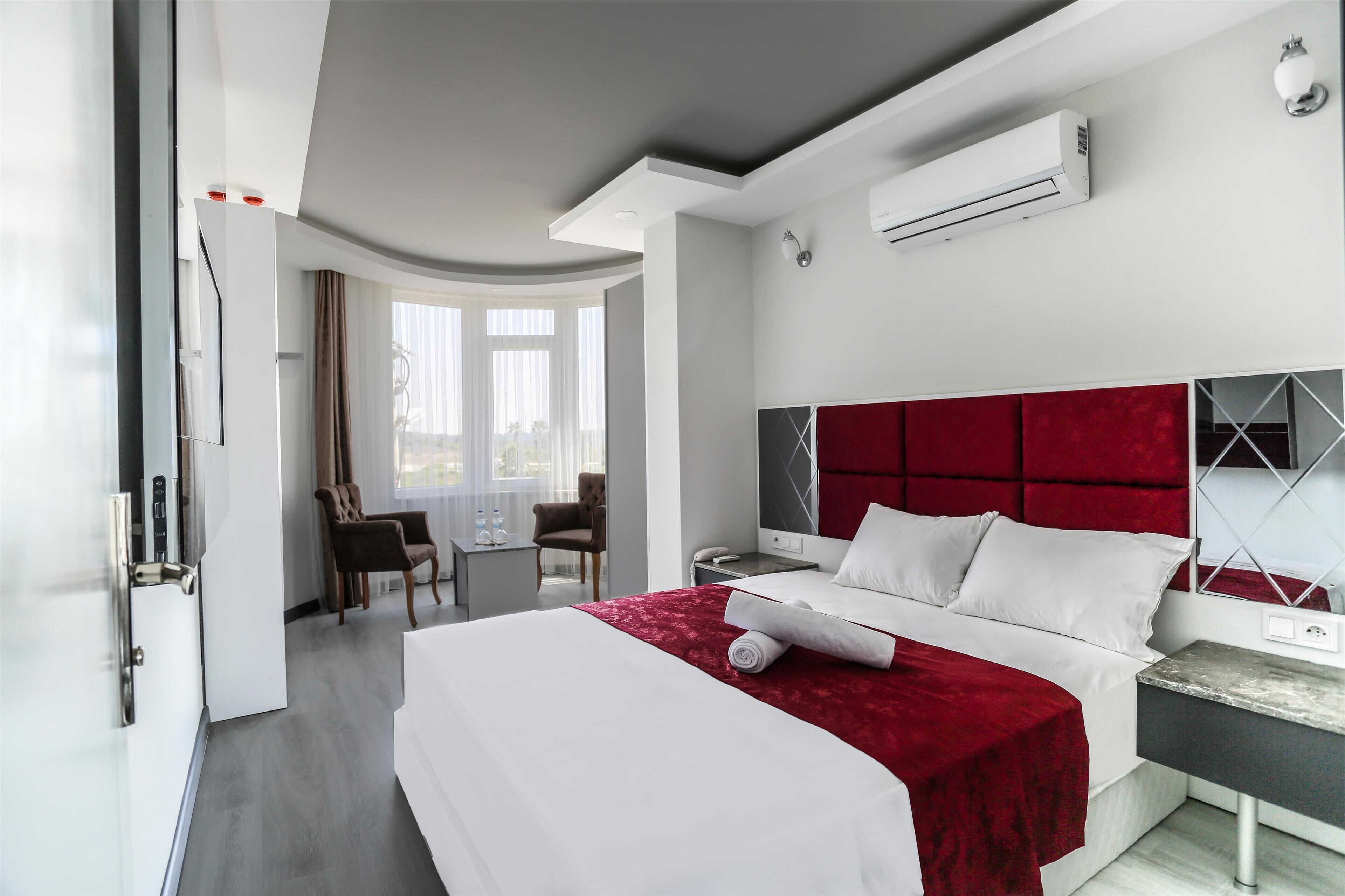 Habitación de hotel con una cama grande, cabecera roja, mesita de noche, escritorio con silla y una ventana con cortinas.