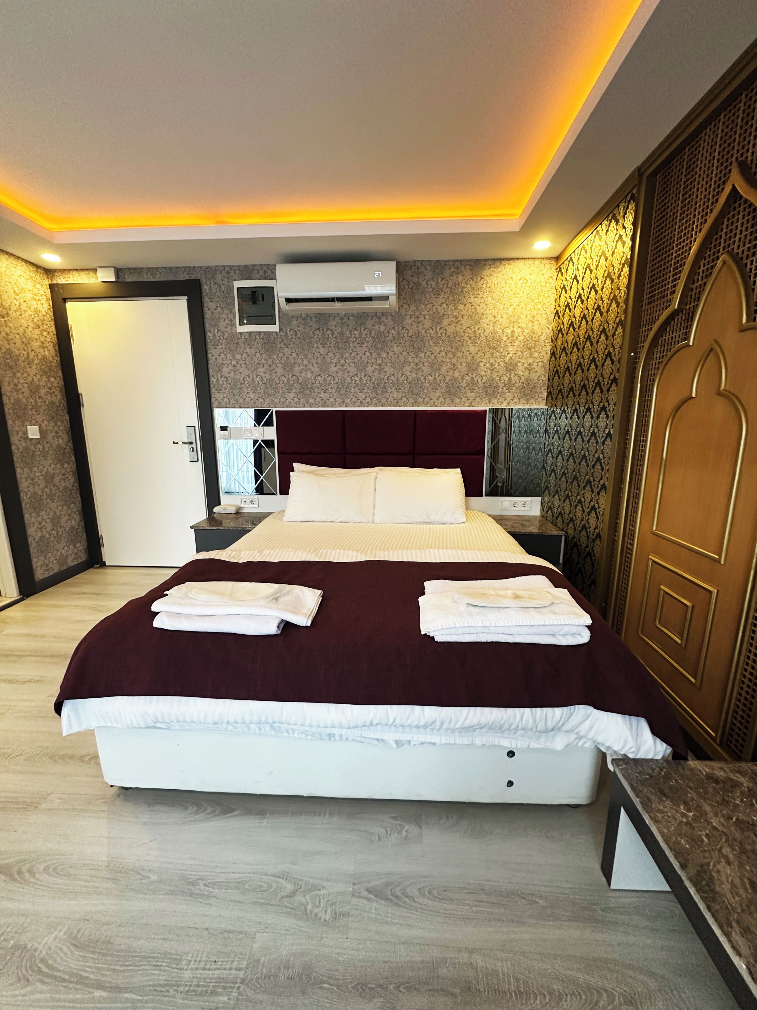 A hotel room with a large bed, a wooden floor, a wall-mounted air conditioner, and a door with a decorative panel.