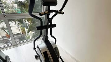 Una bicicleta estática con pantalla digital, ubicada cerca de una ventana con vistas a árboles y edificios.