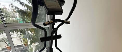 Una bicicleta estática con pantalla digital, ubicada cerca de una ventana con vistas a árboles y edificios.