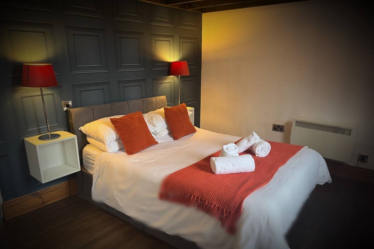 A hotel room with a bed, two nightstands, a red lamp on each, a white blanket, and rolled towels on the bed.