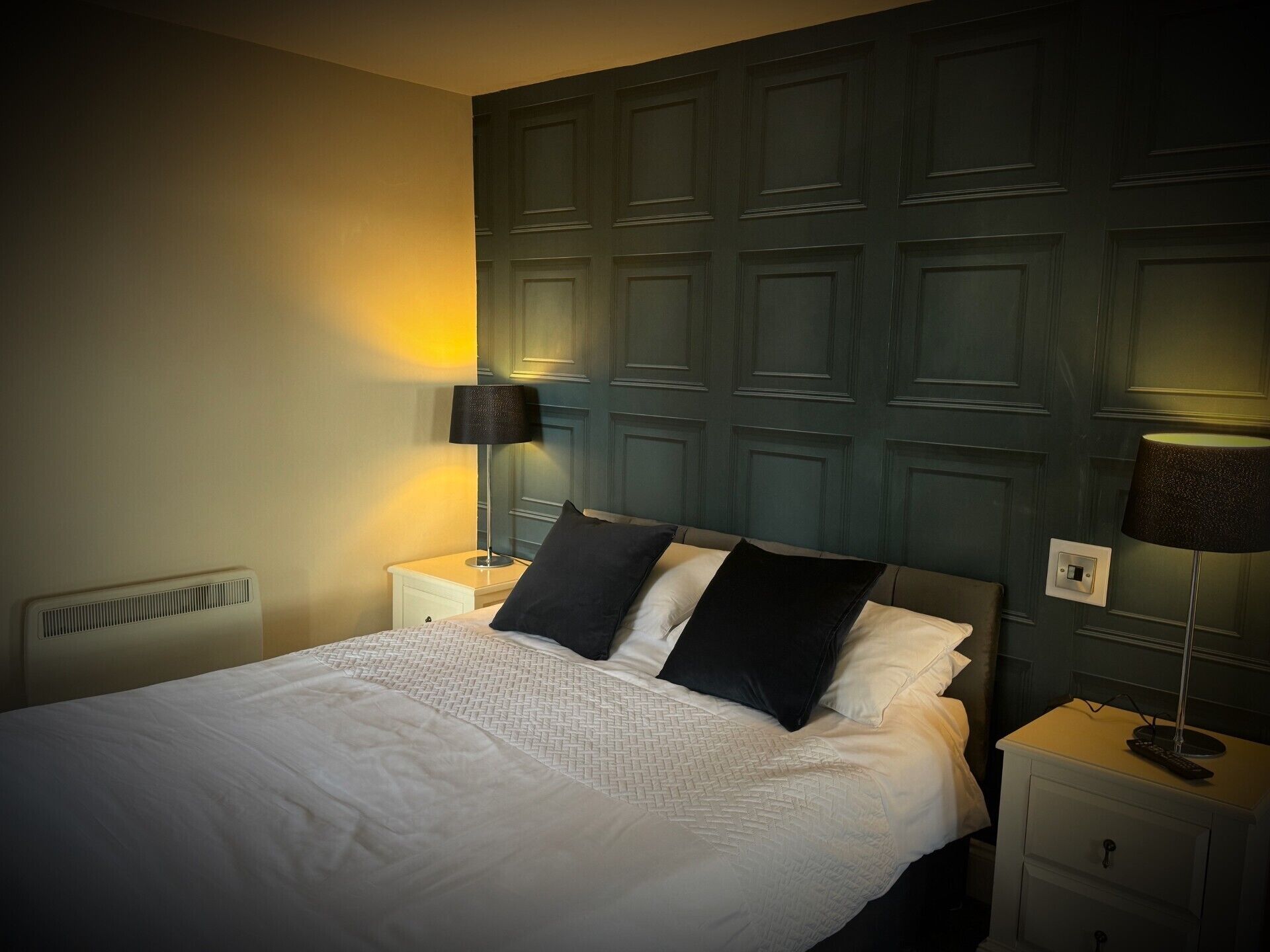 Een netjes opgemaakt bed met witte lakens en zwarte kussens, met aan weerszijden nachtkastjes met lampen, in een kamer met een donker houten hoofdbord.