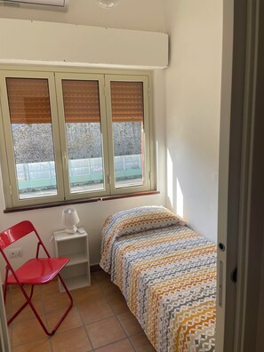 Una stanza modesta con un letto singolo, una sedia rossa, un tavolino con una lampada e una finestra con persiane.