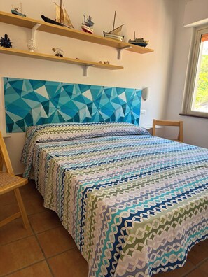 Una camera da letto con una testiera a motivi geometrici, un letto con biancheria a fantasia, una sedia di legno e uno scaffale con modellini di navi.