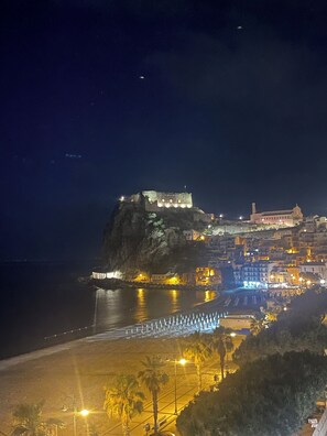 Un pueblo costero con una imponente estructura similar a un castillo en un acantilado, un paseo marítimo y edificios con iluminación cálida.