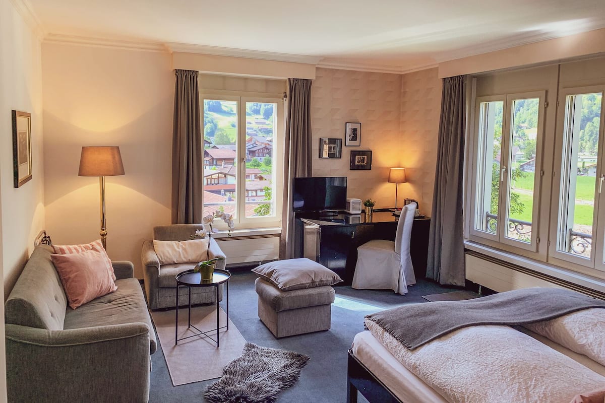 Une chambre d’hôtel avec un canapé, une petite table, un bureau, une chaise et un lit. Il y a des fenêtres avec des rideaux, une télévision et une lampe.