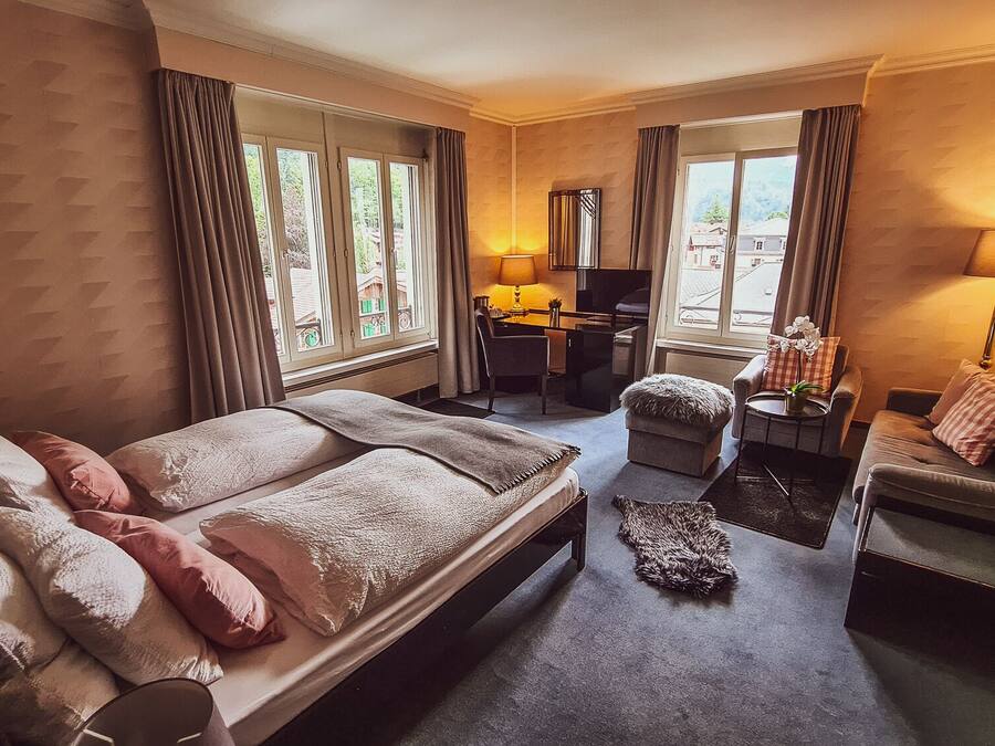 Habitación de hotel con cama, escritorio, silla y sofá. Se ven ventanas con cortinas, una lámpara y una planta.