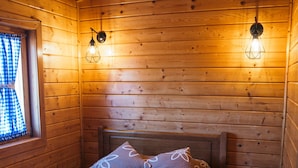 A cozy wooden cabin room with a bed, two wall-mounted lights, a small round table, and a window with blue curtains.