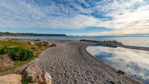 Une plage bordée d’un rivage rocheux, avec une eau calme et des montagnes lointaines se découpant sur un ciel partiellement nuageux.