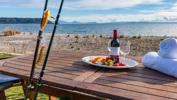 Une table en bois, avec une bouteille de vin, un verre et une assiette de fruits, près d’un lac.