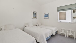 Une chambre avec trois lits simples, du linge de lit blanc, deux tableaux encadrés au mur, une fenêtre avec des stores et deux chaises en plastique blanches.