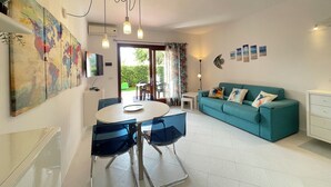 A modern living room with a teal sofa, a dining table with chairs, a large abstract painting, and a flat-screen TV mounted on the wall.