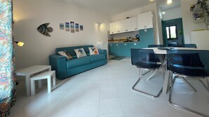 A modern living room with a teal sofa, white coffee table, and black chairs. A kitchen with white cabinets and a blue countertop is visible in the background.