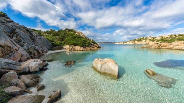 A coastal landscape with clear turquoise waters, rocky shores, and a large boulder in the foreground.