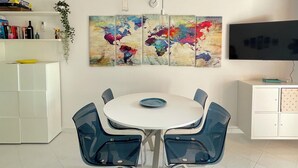 A modern dining area with a white table and blue chairs, a world map artwork above, and a flat-screen TV on a white cabinet.