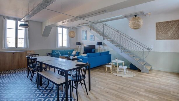 A spacious living area with a blue sofa, a dining table with chairs, and a staircase with a metal railing. - PHILIPPE AUGUSTE LOFT 5 - Loft for 12 - 5 rooms - BASTILLE - PÈRE LACHAISE (Paris)