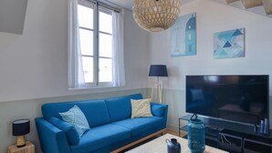 A modern living room with a blue sofa, a flat-screen TV, a coffee table, and a window with curtains. - PHILIPPE AUGUSTE LOFT 5 - Loft for 12 - 5 rooms - BASTILLE - PÈRE LACHAISE (Paris)