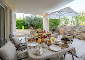 Un área de comedor al aire libre con una mesa lista para el desayuno, que incluye frutas, pasteles y bebidas.