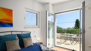 Un dormitorio con cama, almohadas, mesita de noche, lámpara y vista a un balcón exterior con mesa y sillas.
