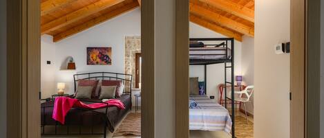 A room with a sloped wooden ceiling, a bed with a pink blanket, a bunk bed, a desk with a computer, and a chair.