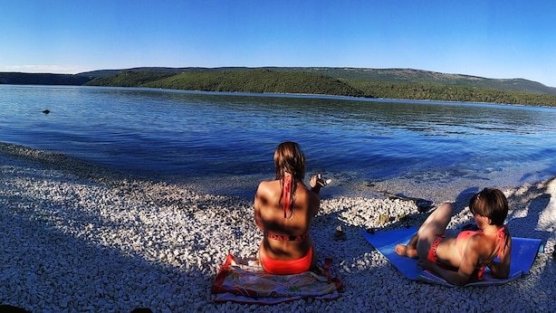 Dos personas en una playa de guijarros con un lago y colinas al fondo.