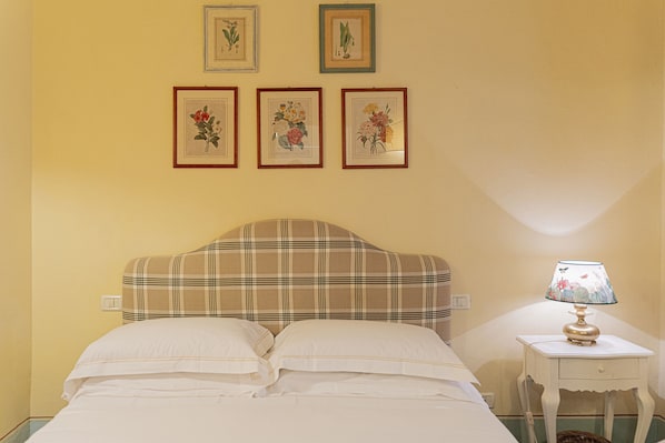 Un dormitorio con una cama, una mesita de noche con una lámpara y cuatro cuadros con motivos florales en la pared.