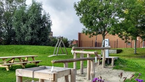 A playground with swings, a swing set, and picnic tables in a grassy area with trees and a building in the background.