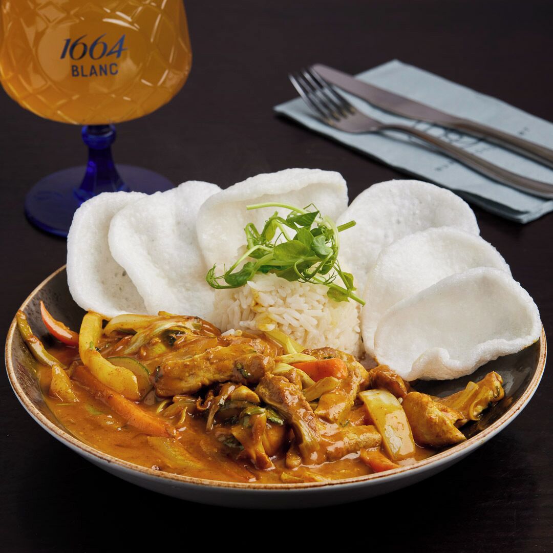 Een bord eten met rijst, groenten en een currygerecht, vergezeld van een glas witte wijn en een vork en mes op een tafel.