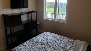 A bedroom with a bed, a TV on a black dresser, a window with a view of a green landscape, and a balcony railing. - Luxurious Apartment with  Wi-Fi and Private Bathroom (Fort McMurray)