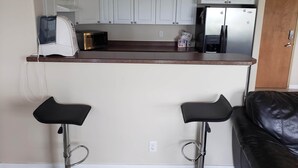 A compact kitchen with white cabinets, a microwave, a toaster, and a refrigerator. There are two black bar stools and a black leather couch in the adjacent living area. - Luxurious Apartment with  Wi-Fi and Private Bathroom (Fort McMurray)