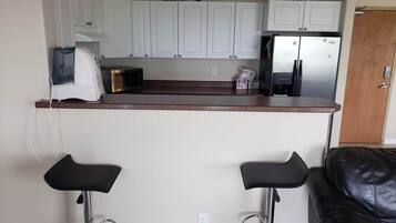 A compact kitchen with white cabinets, a microwave, a toaster, and a refrigerator. There are two black bar stools and a black leather couch in the adjacent living area.