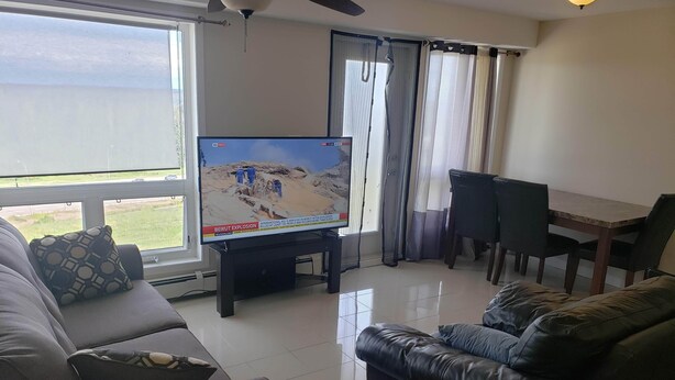 A living room with a television displaying a news broadcast, a sofa, a leather armchair, a dining table with chairs, and a view through the window. - Luxurious Apartment with  Wi-Fi and Private Bathroom (Fort McMurray)
