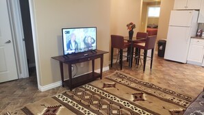 Una sala de estar con televisor, una mesa de comedor con sillas, un tapete estampado y una zona de cocina con refrigerador.