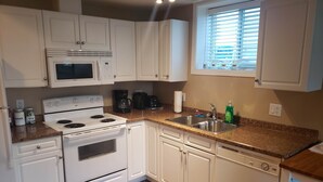 Una cocina con armarios blancos, estufa, microondas, lavabo y ventana con persianas.