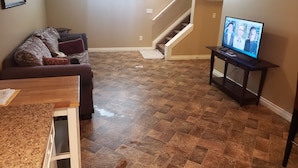 Un salón con suelo de parqué encalado marrón, un sofá marrón con cojines, una mesa de centro de madera, un televisor sobre un mueble bajo y una escalera que sube a un nivel superior.