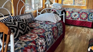 A cozy cabin room with two beds, wooden floors, large windows, and a ceiling fan.