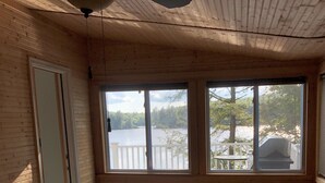 A room with wooden walls and ceiling, a ceiling fan, a window with a view of a lake, a wooden table with a chair, and a plaid sofa.