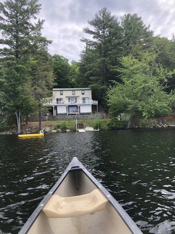 Una canoa en un lago tranquilo, con una casa y árboles al fondo.