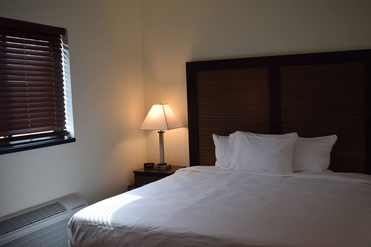 A hotel room with a bed, a nightstand with a lamp, a window with blinds, and a wall-mounted heater.