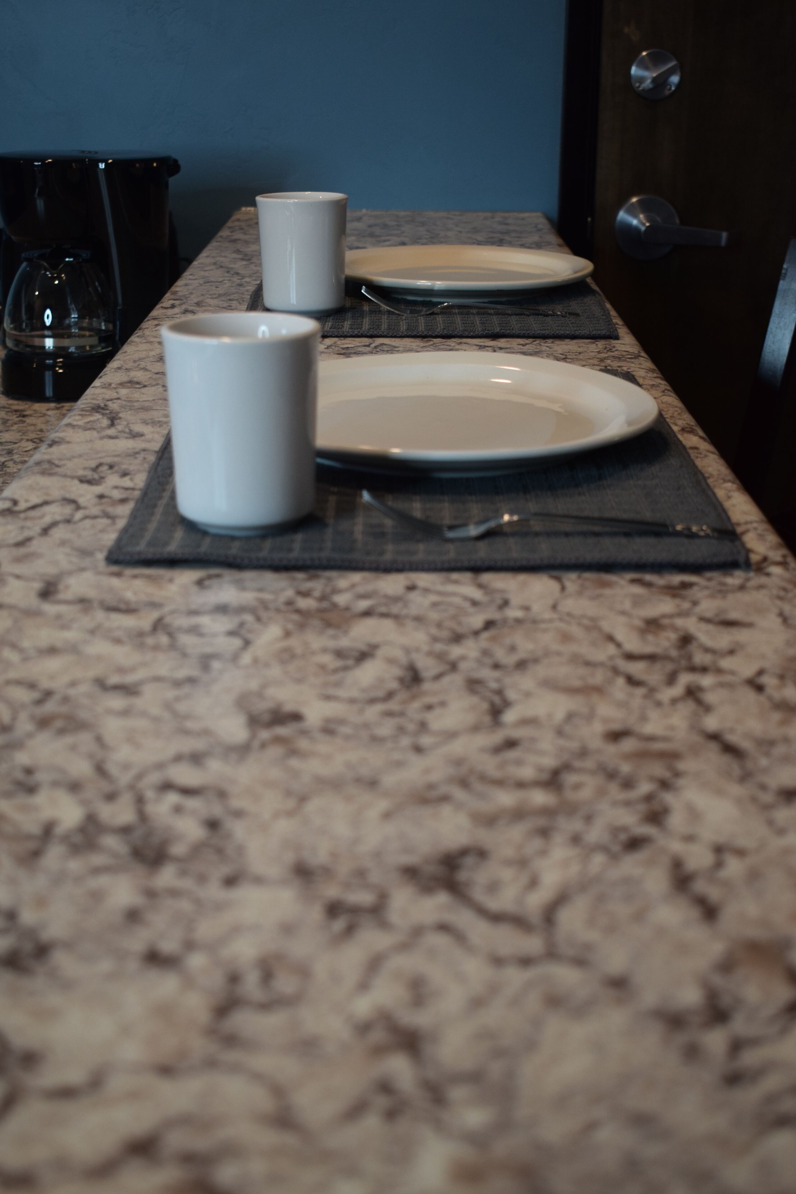 A kitchen counter with a coffee maker, two white mugs, and a plate on a placemat.