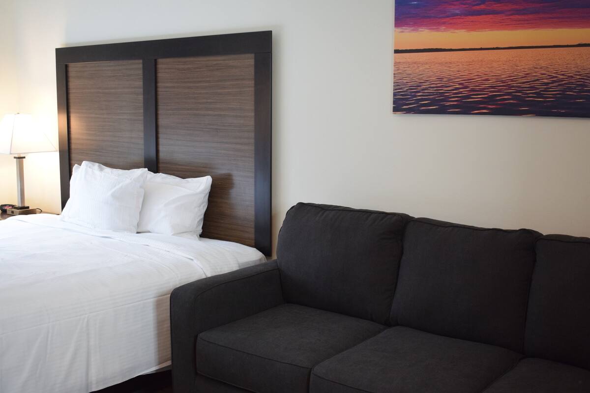 A hotel room with a bed, a dark gray sofa, and a painting of a sunset over water on the wall.