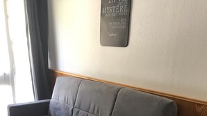 Un sofá gris con estructura de madera, un taburete blanco pequeño y un letrero fijado a la pared con texto en francés.