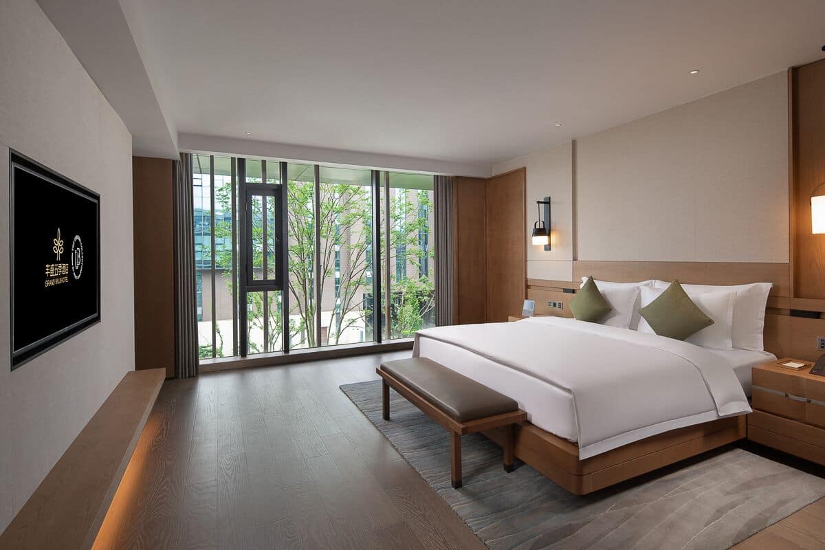 Habitación de hotel moderna con una cama grande, un banco, una mesita de noche, televisión y vistas a zonas verdes por las ventanas.
