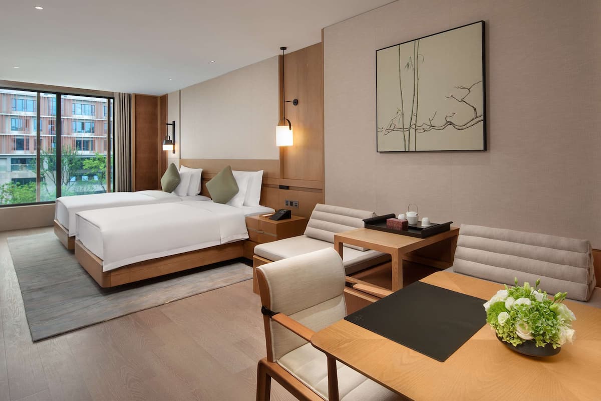 Una habitación de hotel moderna con dos camas, un sofá, una mesa de comedor y un amplio ventanal con vista a la ciudad.
