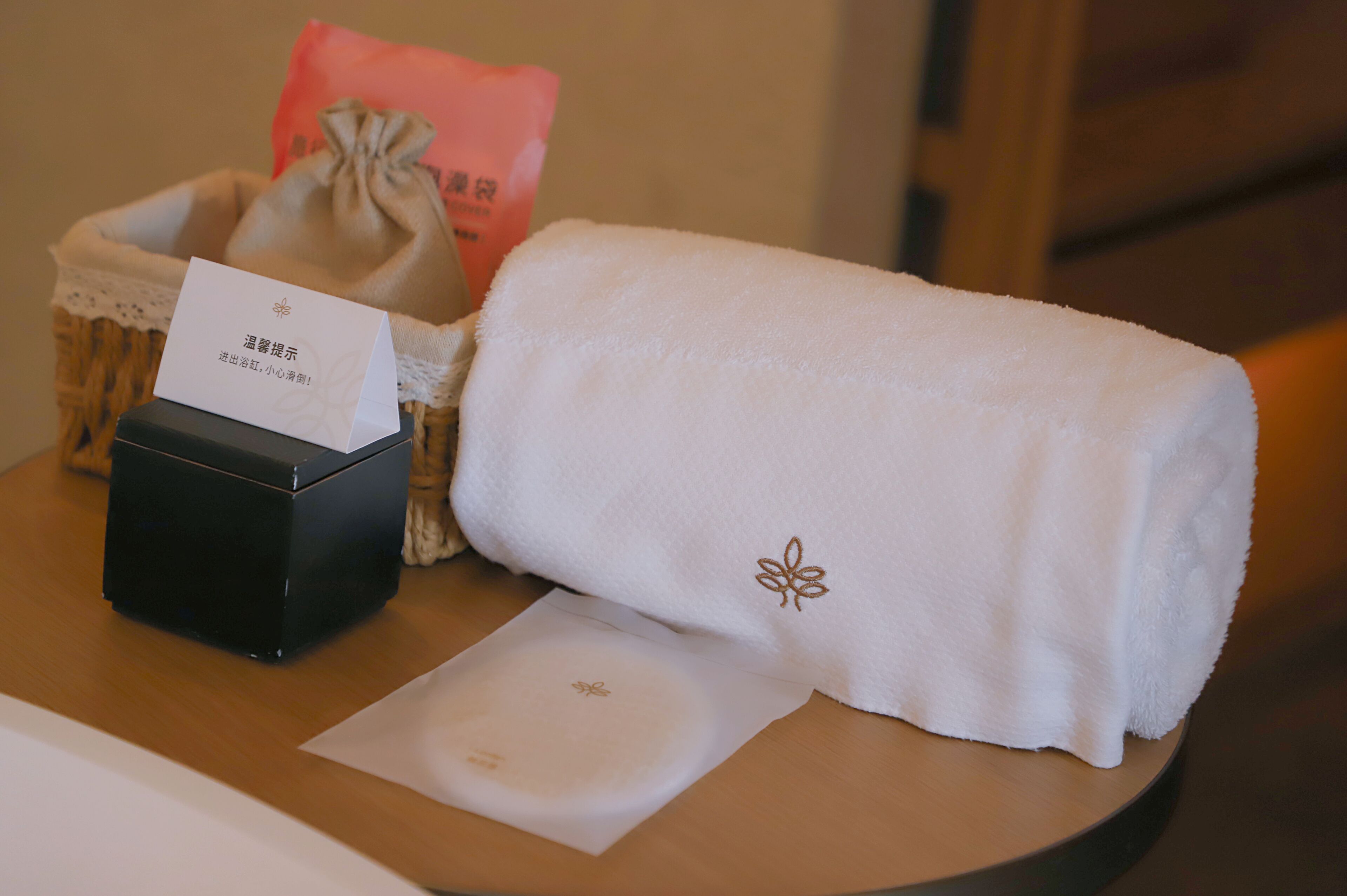 Una caja negra pequeña, una toalla blanca con diseño floral, un bolso rosa y una tarjeta blanca sobre una mesa de madera.
