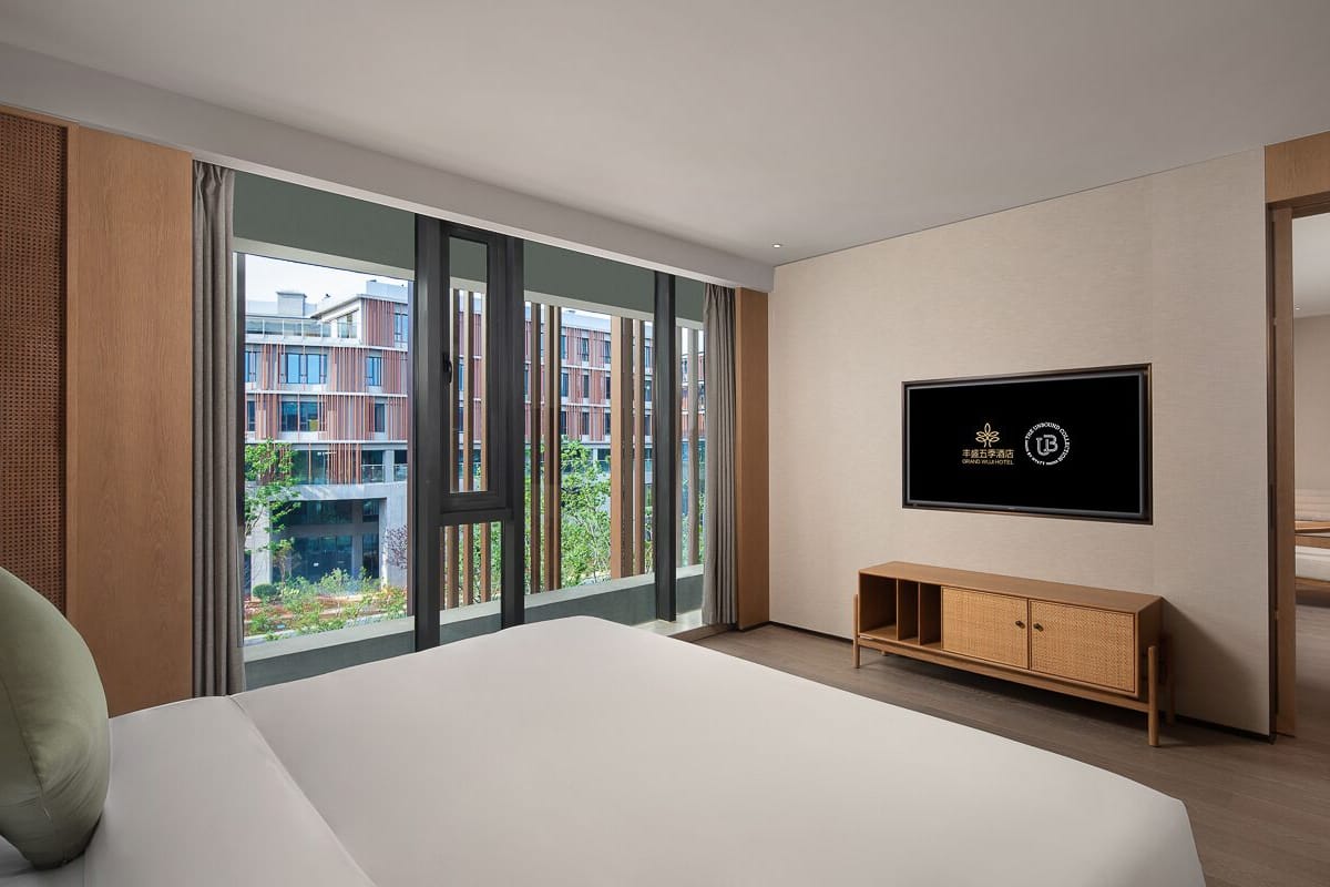 Una habitación de hotel moderna con una cama grande, un televisor montado en la pared y vistas de la ciudad a través de amplias ventanas.