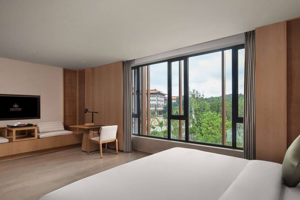 Habitación de hotel con una cama grande, un escritorio con una silla, un televisor y un ventanal con vistas a edificios y árboles.