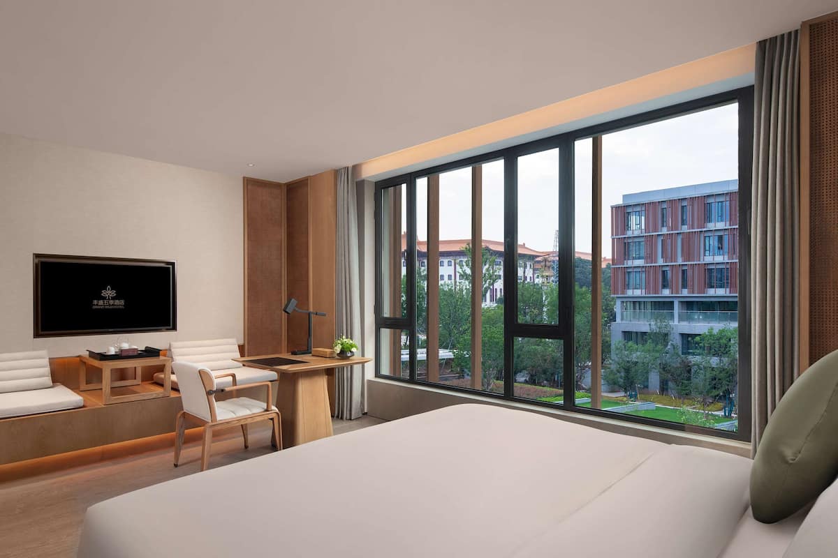 Una habitación de hotel moderna con una cama grande, un escritorio con silla, un televisor montado en la pared y un ventanal con vistas a edificios y árboles.